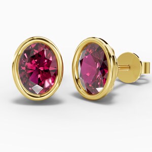 May include: A pair of oval-shaped stud earrings featuring faceted deep pink gemstones set in polished yellow gold bezels. Each earring includes a matching yellow gold post and a secure butterfly-style backing, showcasing a clean, metallic finish.