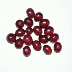 Puede incluir: Un conjunto de gemas ovaladas, pulidas, de color rojo intenso. Las piedras están dispuestas sobre una superficie blanca, mostrando sus superficies lisas y reflectantes. Las gemas varían ligeramente en tamaño y tono, con un acabado brillante.