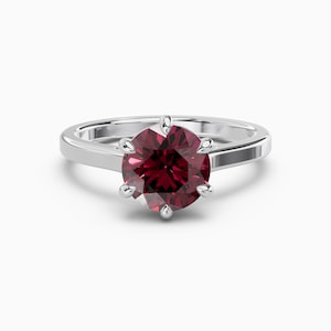 Peut inclure: Une bague en argent avec une grosse pierre précieuse ronde et rouge foncé. La pierre est maintenue par six griffes. Le corps de la bague est lisse et poli. La bague est posée sur fond blanc.