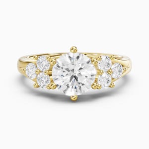 May include: A yellow gold ring featuring a large, round, brilliant-cut centre stone held by four prongs. The band is accented on each side by a cluster of three smaller, round, clear stones, creating a symmetrical, multi-stone design.