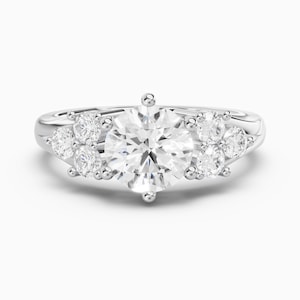 May include: A silver-coloured ring featuring a large, round, brilliant-cut centre stone flanked by three smaller round stones on each side, all set in a polished metal band that tapers slightly towards the back of the ring.