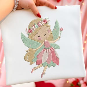Op de afbeelding: Wit vierkant kussen met een geborduurd feeënontwerp. De fee heeft blond haar, een bloemenkrans en groene en roze vleugels. Ze houdt een sterrenvormige toverstaf vast en draagt een roze jurk.