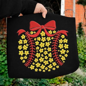 Puede incluir: Bolso tote negro con un diseño floral. El diseño incluye un lazo y una cinta rojos, con una disposición central de flores amarillas. El bolso lo sostiene una persona, con el fondo borroso.