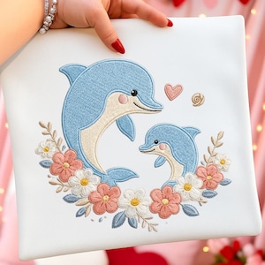 Könnte beinhalten: Quadratischer weißer Stoff mit einem gestickten Design von zwei blauen Delfinen, umgeben von einem Blumenkranz aus rosa, weißen und gelben Blumen. Ein rosa Herz und eine Spirale sind ebenfalls gestickt.