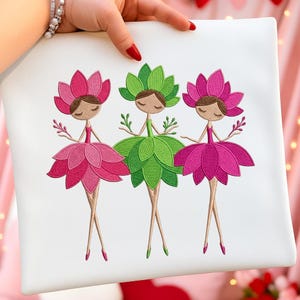 以下が含まれることがあります： 白い正方形のクッション。3人の花の妖精の刺繍デザイン。各妖精は、ピンクと緑の色合いの花の形をしたヘッドピースとスカートを持っています。妖精たちは腕を広げています。