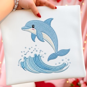 May include: White square pillow with an embroidered design of a blue dolphin leaping over a wave. The dolphin has a light pink mouth and belly. The wave and dolphin are detailed with light blue and white stitching.