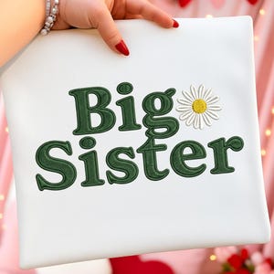 May include: White pillowcase with the embroidered text "Big Sister" in dark green, accompanied by a daisy. The pillowcase is held by a hand with red nail polish. The design is simple and elegant, perfect for a new big sister.