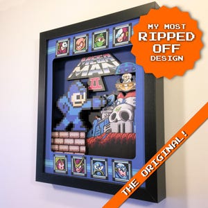 May include: A framed 3D artwork of the video game character Mega Man. The black framed artwork includes the text "MY MOST RIPPED OFF DESIGN" and "THE ORIGINAL!". The artwork is in a pixelated style with a blue background.