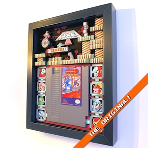 May include: Framed artwork featuring a Mega Man 2 NES cartridge. The black frame surrounds a 3D diorama of the game's levels and characters. The artwork includes the text "THE ORIGINAL!" on an orange diagonal stripe.