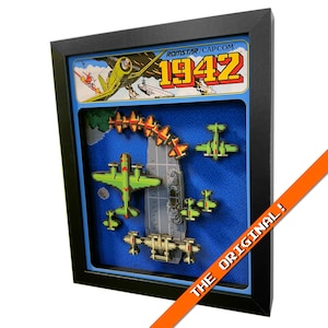 Peut inclure: Œuvre d'art 3D encadrée représentant une scène de bataille d'avions pixélisée du jeu vidéo 1942. L'œuvre comprend des avions verts et rouges, un porte-avions gris et le logo du jeu. Le cadre est noir et le fond est bleu.