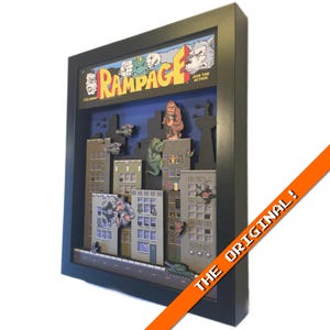 May include: Framed artwork featuring a 3D diorama of the classic arcade game "Rampage." The scene includes skyscrapers, monsters, and the game's logo. The frame is black, and an orange banner reads "THE ORIGINAL!"