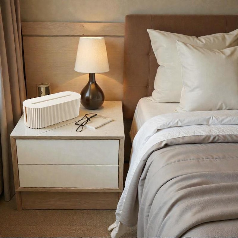 K&ouml;nnte beinhalten: Eine Schlafzimmerszene mit einem Nachttisch mit einem cremefarbenen Kabelorganizer, einer braunen Lampe und einem Bett mit wei&szlig;er und beiger Bettw&auml;sche. Der Nachttisch hat zwei Schubladen und eine Marmorplatte. Das Bett hat ein braunes Kopfteil.