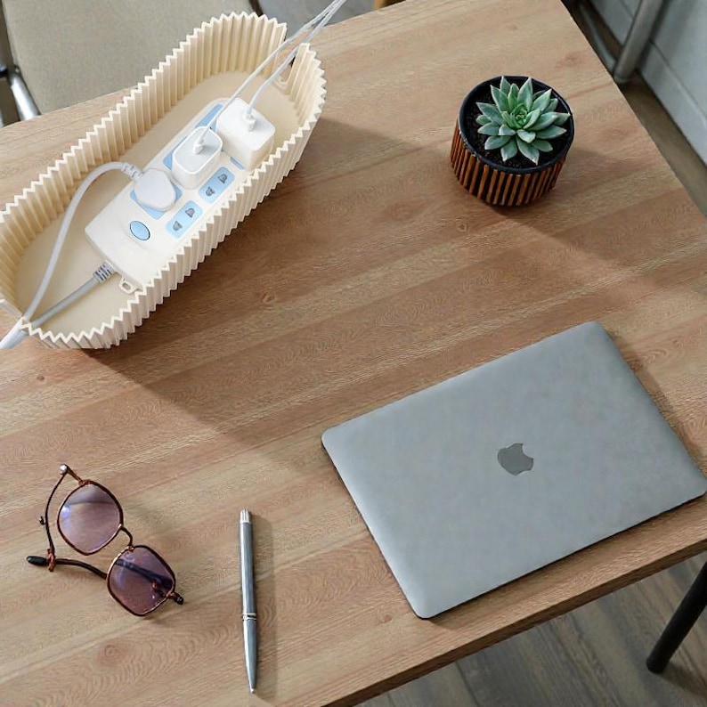 K&ouml;nnte beinhalten: Ein helles Holzschreibtisch mit einer beigen Steckdosenleiste, einem geschlossenen silbernen Laptop, einer kleinen Topf-Sukkulente, einer Brille und einem Stift. Die Steckdosenleiste befindet sich in einem beigen Beh&auml;lter mit gewelltem Rand.