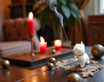 May include: A white ceramic cat incense holder with a red collar and gold bell holds a lit incense stick, with smoke rising. The cat sits on a wooden table with gold spheres, candles, and a decorative background.