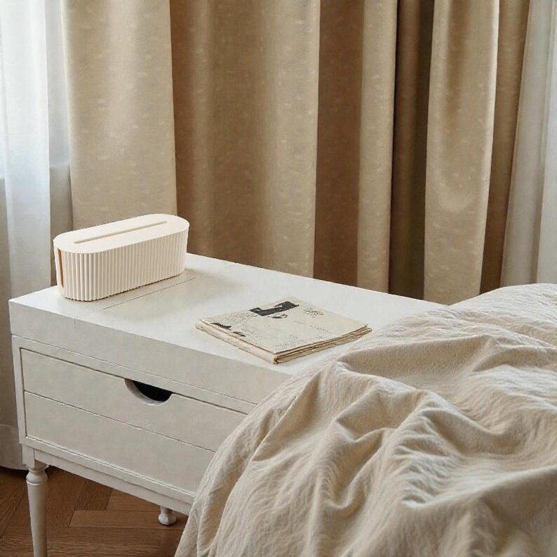 K&ouml;nnte beinhalten: Eine cremefarbene Taschentuchbox mit vertikalen Rippendetails steht auf einem wei&szlig;en Nachttisch. Eine Zeitung liegt daneben. Die Szene spielt vor beigefarbenen Vorh&auml;ngen und einem Bett mit einer cremefarbenen Bettdecke.