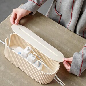 K&ouml;nnte beinhalten: Ein cremefarbener Kabelorganizer mit geripptem Design, der mehrere wei&szlig;e Netzadapter und Kabel enth&auml;lt. Der Deckel wird angehoben und gibt den Inhalt frei. Der Organizer dient dazu, Kabel zu verbergen und zu verwalten.