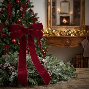 May include: A large, deep red velvet bow with two long, flowing tails. The bow features a structured centre knot and smooth, soft-textured fabric. It is positioned in the foreground, showcasing a rich, dark crimson colour and a classic shape.