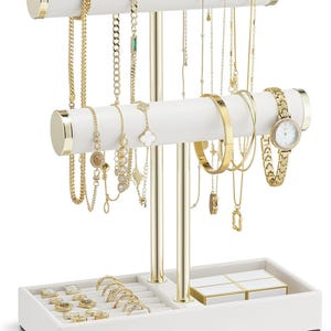 Op de afbeelding: Een wit en goudkleurig sieradenrek met diverse gouden kettingen, armbanden, ringen en een horloge. De organizer heeft twee witte staven met gouden accenten om kettingen op te hangen, een bakje voor ringen en een klein doosje.