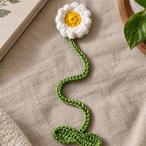 May include: A handmade crochet bookmark shaped like a daisy flower. The flower head features white petals surrounding a bright yellow center. A long, wavy green stem extends downward, ending in two small, textured green leaves at the base.
