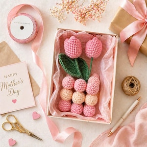 Puede incluir: Una caja de regalo con un arreglo de tulipanes rosas de ganchillo, hojas verdes y capullos color melocotón. La caja está rodeada de cinta rosa, una tarjeta de "Happy Mother's Day", tijeras doradas y un regalo envuelto.