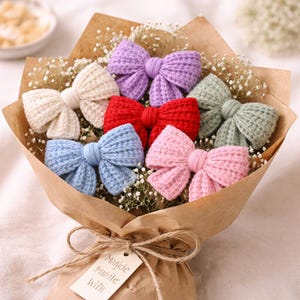May include: A bouquet of colourful, knitted bows in shades of cream, purple, red, blue, pink, and green. The bows are presented with baby's breath, wrapped in brown paper, and tied with twine. A tag reads "Manide & made with".