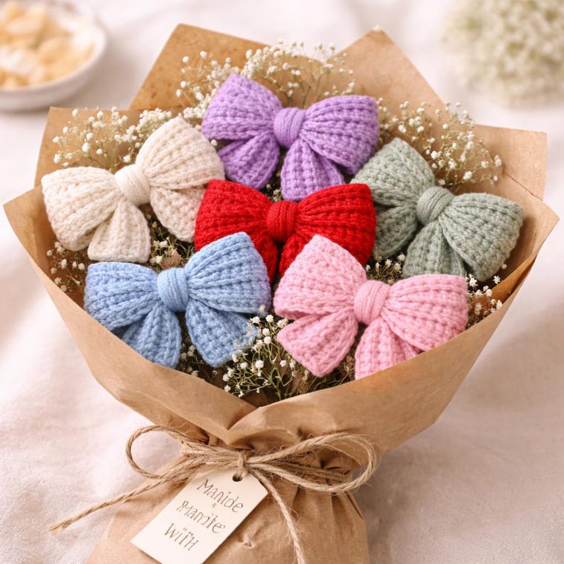 May include: A bouquet of colourful, knitted bows in shades of cream, purple, red, blue, pink, and green. The bows are presented with baby's breath, wrapped in brown paper, and tied with twine. A tag reads "Manide & made with".