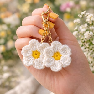 May include: Two white crochet daisy flower keyrings with yellow centres, attached to a gold-coloured clasp and chain. The keyrings are held up against a blurred background of flowers.