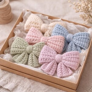 May include: A selection of pastel-coloured, handmade crochet bows, including pink, blue, green, and cream. The bows are presented in a brown cardboard box, nestled on white tissue paper. These bows are a delightful accessory.