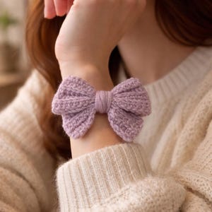 May include: A lavender knitted bow bracelet. The bow is centered and has a textured appearance. The bracelet is worn on the wrist. The background is a cream-coloured sweater.
