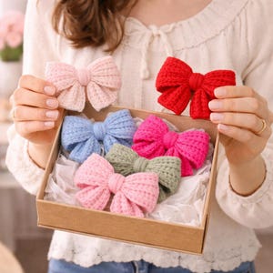 May include: A selection of knitted bows in a range of colours: pink, red, blue, and green. The bows are presented in a cardboard box with white tissue paper. One pink bow is held up separately.