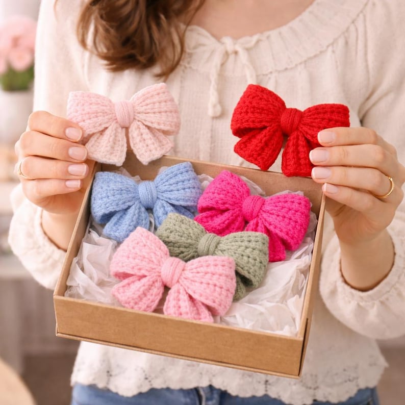 May include: A selection of knitted bows in a range of colours: pink, red, blue, and green. The bows are presented in a cardboard box with white tissue paper. One pink bow is held up separately.