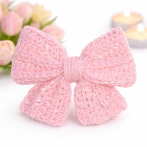 May include: A light pink, hand-crocheted bow. The bow is knotted in the middle and has a textured finish. The background features soft pink flowers and two lit candles, creating a delicate and feminine aesthetic.