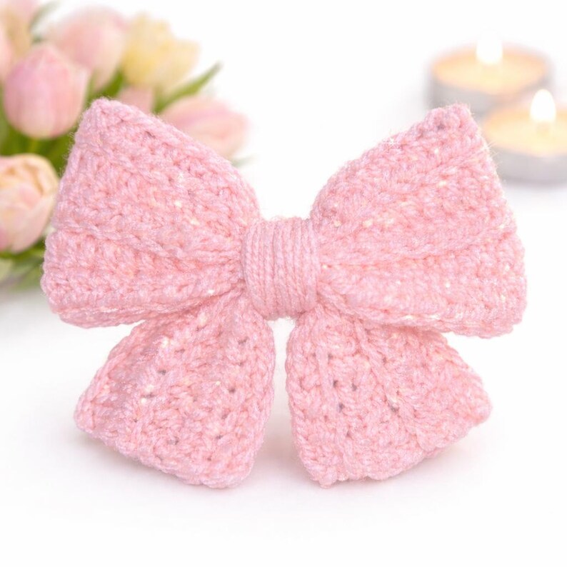 May include: A light pink, hand-crocheted bow. The bow is knotted in the middle and has a textured finish. The background features soft pink flowers and two lit candles, creating a delicate and feminine aesthetic.