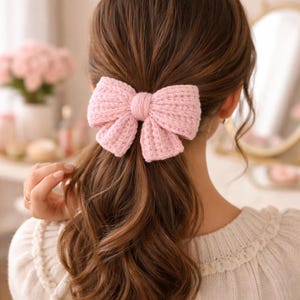 May include: A pale pink knitted bow hair accessory. The bow is tied in the centre and attached to a hair clip. The hair accessory is worn in the hair, with brown hair flowing down the back.