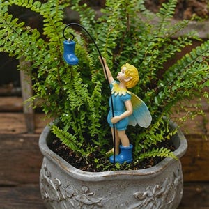 May include: A miniature fairy figurine in a blue outfit with yellow hair and wings, holding a fishing rod with a blue boot attached. The fairy is placed in a decorative gray flower pot filled with green plants.