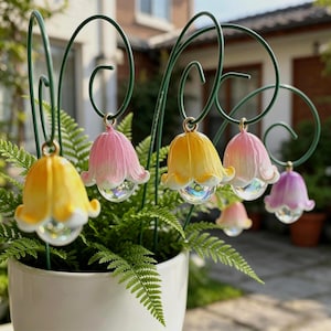 Könnte beinhalten: Eine Sammlung farbenfroher, glockenförmiger Gartenornamente. Jedes Ornament hängt an einem grünen Metallstab und hat eine durchscheinende, kugelförmige Basis. Die Ornamente sind in Gelb-, Rosa- und Lilatönen gehalten und verleihen dem Garten einen verspielten Touch.