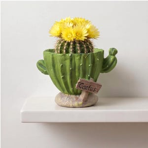 Pode incluir: Um vaso verde em forma de cacto com uma pequena placa castanha com a palavra Cactus escrita a preto. O vaso contém um cacto verde redondo com um grupo de flores amarelas brilhantes. A base do vaso tem um acabamento texturado de pedra.