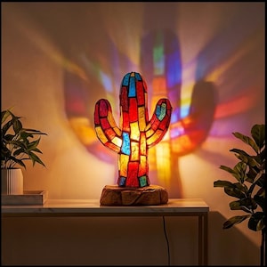 May include: A stained glass cactus lamp featuring a mosaic of red, yellow, and blue glass pieces. The lamp sits on a textured, rock-like base and casts a vibrant, multi-colored light projection onto the wall behind it, creating a glowing effect.