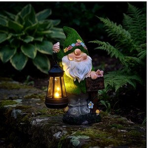 Può includere: Una statuetta di gnomo da giardino con un cappello verde decorato da una corona di fiori e una tunica verde. Lo gnomo ha una lunga barba bianca, tiene una lanterna nera che emette una luce gialla calda e poggia la mano su un cartello Welcome to my Garden.