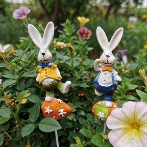 Può includere: Due picchetti da giardino a forma di conigli bianchi antropomorfi. Uno indossa un gilet giallo con papillon blu e pantaloni verdi su una base arancione. L'altro indossa una camicia bianca con salopette blu e un papillon di colore arancione.