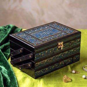 May include: A rectangular dark wood jewelry box with a tiered accordion-style opening mechanism. The lid and sides feature intricate blue and gold painted patterns. A small gold-toned metal latch secures the front of the box.