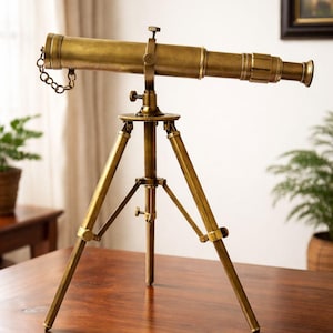 May include: A brass telescope on a tripod, showcasing a vintage aesthetic. The telescope's barrel is cylindrical, with a chain attached. The tripod has three legs, and the entire piece is set on a wooden surface. The brass finish gives it a classic, antique look.