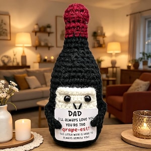 May include: A handcrafted crochet wine bottle cover in black and claret, featuring a white face and black button eyes. It holds a sign that reads "DAD I'll always love you, you're the grape-est! This little wine is here to always remind you!"