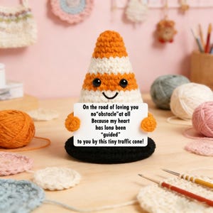 May include: A handmade orange and white traffic cone with a smiling face and a black base. The cone holds a sign that reads "On the road of loving you no obstacle at all..." This crochet item is a unique gift.