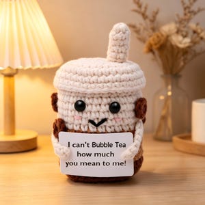May include: A crocheted bubble tea plushie with a cream-colored top and a brown base. The plushie has black eyes, a smiling face, and holds a sign that reads, "I can't Bubble Tea how much you mean to me!"