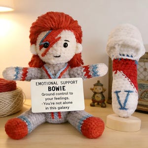 May include: A handmade crochet doll resembling David Bowie, with red hair and a lightning bolt on its face. The doll is wearing a gray and red outfit and holds a card that reads "EMOTIONAL SUPPORT BOWIE". A matching microphone is also present.