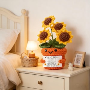 May include: A crocheted sunflower arrangement in an orange pot with a smiling face. The sunflowers are yellow with brown centers. A white card on the pot reads "TO MY MOM" with a loving message. The arrangement is on a white nightstand.