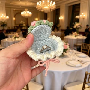 Könnte beinhalten: Eine gehäkelte Ringbox in Form einer Muschel, in Hellblau, Weiß, Grün und Rosa. Die Box enthält einen silbernen Ring mit einem Diamanten. Die Box ist mit einer rosa Schleife gebunden und mit kleinen weißen Perlen verziert.