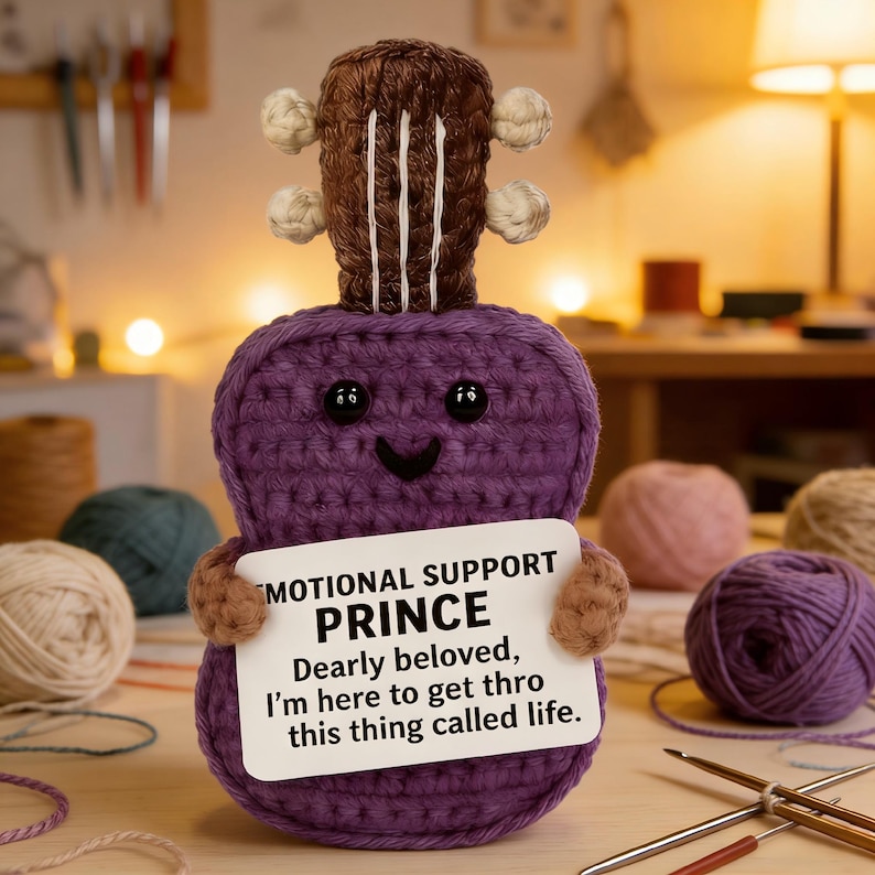 May include: A handmade, purple crochet ukulele with a smiling face and a brown headstock. It holds a sign that reads "MOTIONAL SUPPORT PRINCE, Dearly beloved, I'm here to get thro this thing called life." The ukulele has black button eyes.