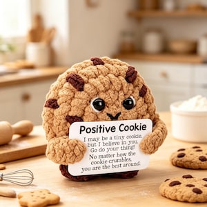 Op de afbeelding: Een rond, beige, gehaakt koekje als knuffel met donkerbruine chocoladestukjes, grote zwarte ogen en kleine armpjes die een wit rechthoekig bordje vasthouden met de tekst: Positive Cookie, I may be a tiny cookie, but I believe in you. Go do your thing!